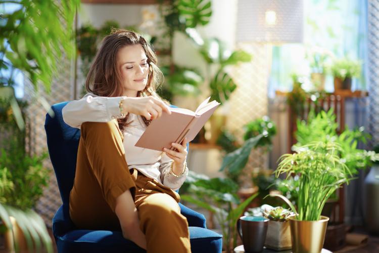 Žena sedi u fotelji i čita svoju knjigu okružena zelenim biljkama